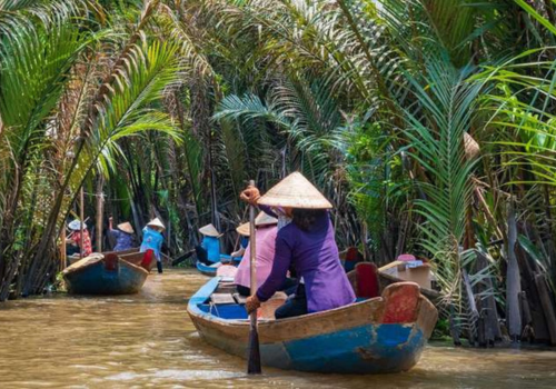Rowing boat on the canals of the Mekong River, surrounded by lush tropical vegetation with tourists wearing traditional Vietnamese conical hats.